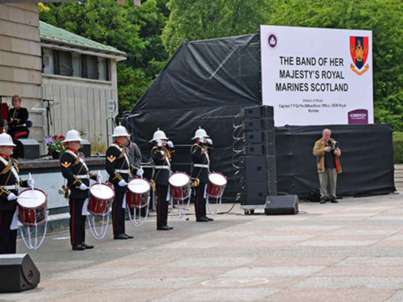 Banda de la Reina de los Marines de Escocia en un desfile militar, con músicos con uniformes militares y instrumentos musicales en una plaza al aire libre.