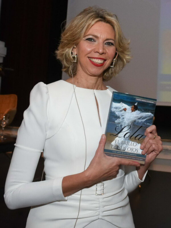Una mujer en un evento de presentación de libro, sosteniendo un libro titulado 'El brillo de sus ojos' en una elegante vestimenta blanca.