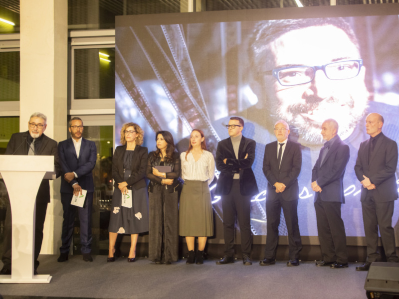 Una celebración formal con personas en trajes y una pantalla proyectando una imagen de un hombre con barba y gafas.