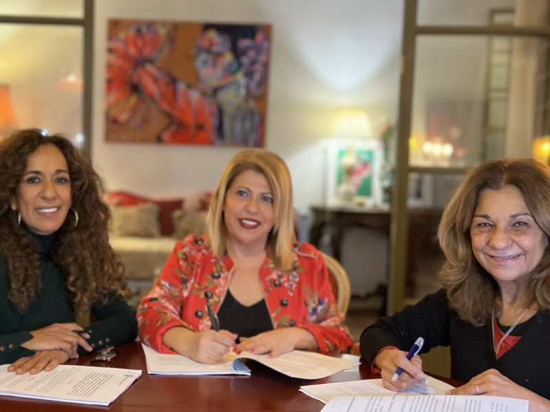 Tres mujeres sentadas en una mesa firmando documentos en un ambiente acogedor con arte en la pared.