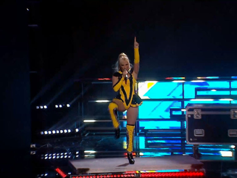 Artista en escenario con ropa brillante y luces de fondo azules, actuando con energía.