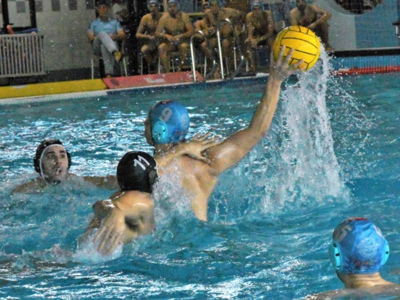 Juego de waterpolo en acción, con jugadores en la piscina y un balón amarillo en el aire. El agua es azul y hay espectadores en la orilla. El agua está salpicada por el movimiento de los jugadores.