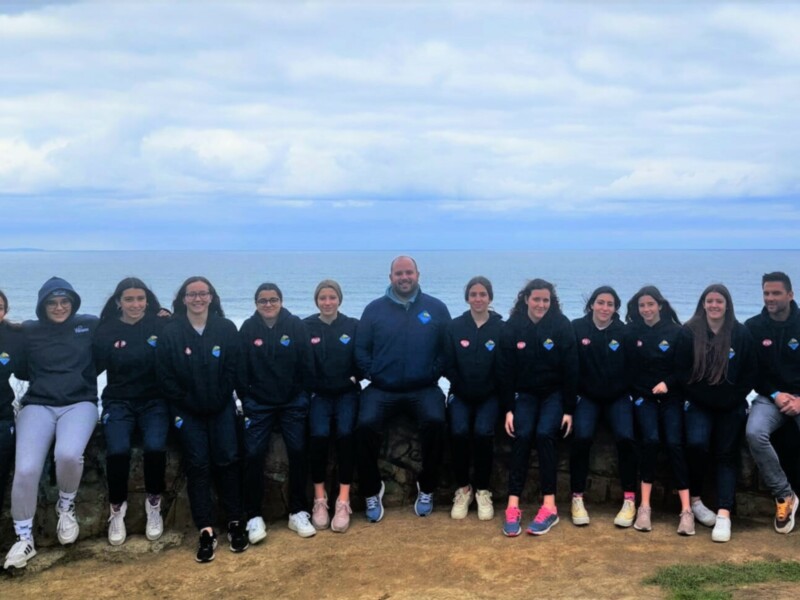 Equipo de jóvenes y adultos posando juntos en una roca costera con el mar y el cielo nublado de fondo.