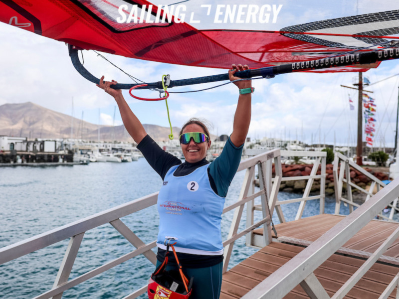 Una mujer sonríe mientras sostiene un par de remos de vela en un puerto deportivo, con montañas en el fondo y banderas ondeando en el horizonte.