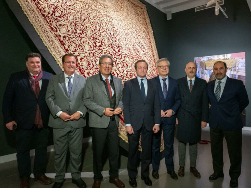 Grupo de hombres en trajes posando frente a una alfombra persa en un espacio interior.