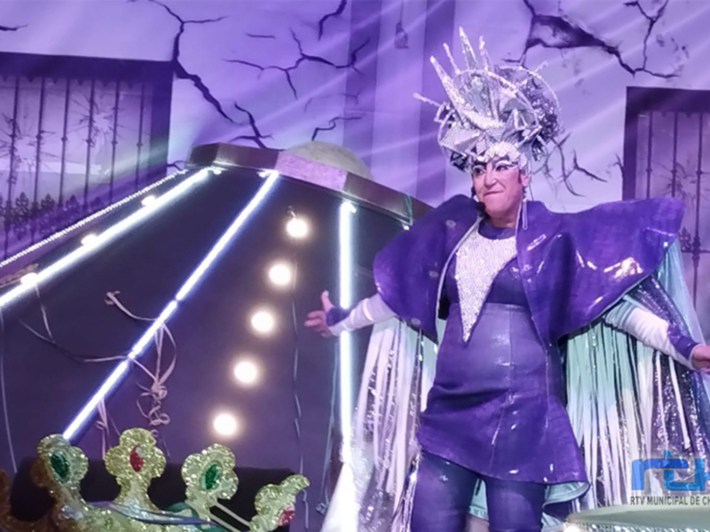 Una figura vestida con un traje púrpura brillante y una corona de plata, posa con las manos extendidas en un escenario iluminado con luces de neón y una decoración de fondo.