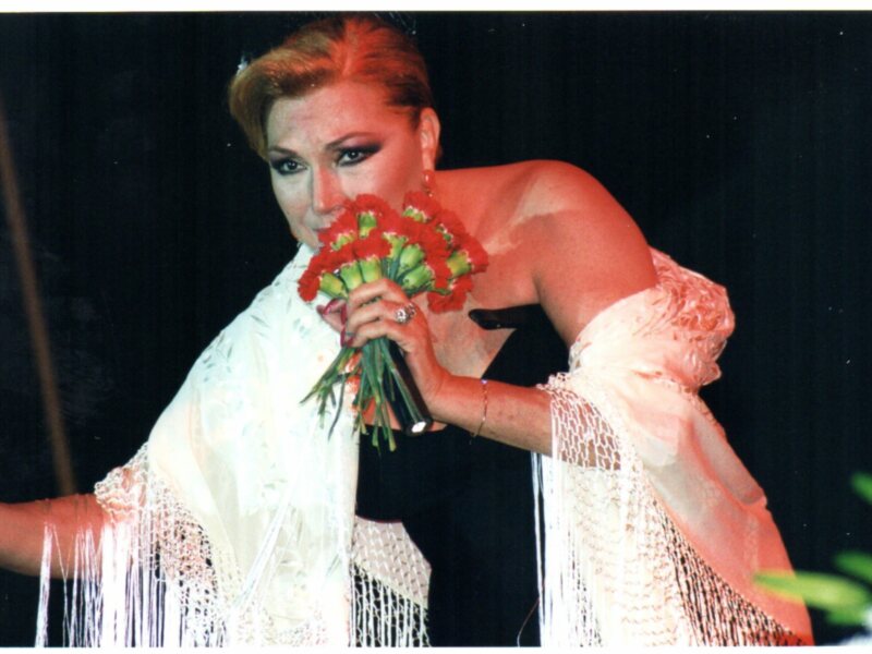 Actriz con ramo de flores rojas, vestida con traje blanco y negro, posando en escenario.