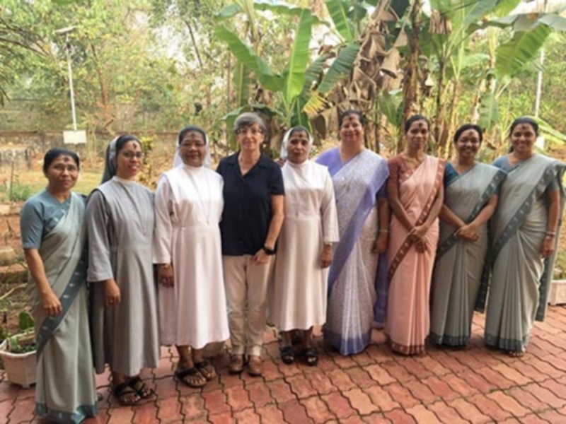 Un grupo de mujeres vestidas con saris y túnicas posa juntas en un entorno tropical con plantas de banano al fondo.