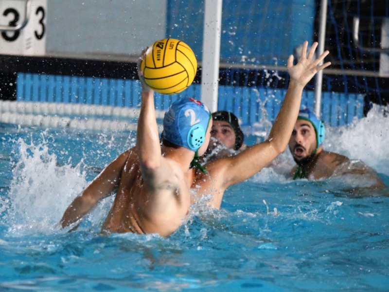 Jugadores de waterpolo en acción durante un partido en una piscina azul. El jugador en el centro sostiene la pelota amarilla, mientras los otros dos están en la acción de la jugada.