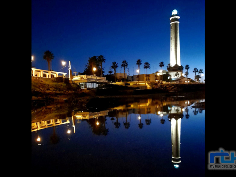 Luminoso faro nocturno reflejado en el agua, con palmeras y luces de la ciudad en el fondo.