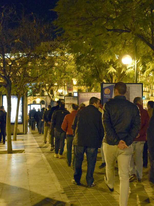Gente caminando por una calle nocturna con luces de árboles y carteles publicitarios.