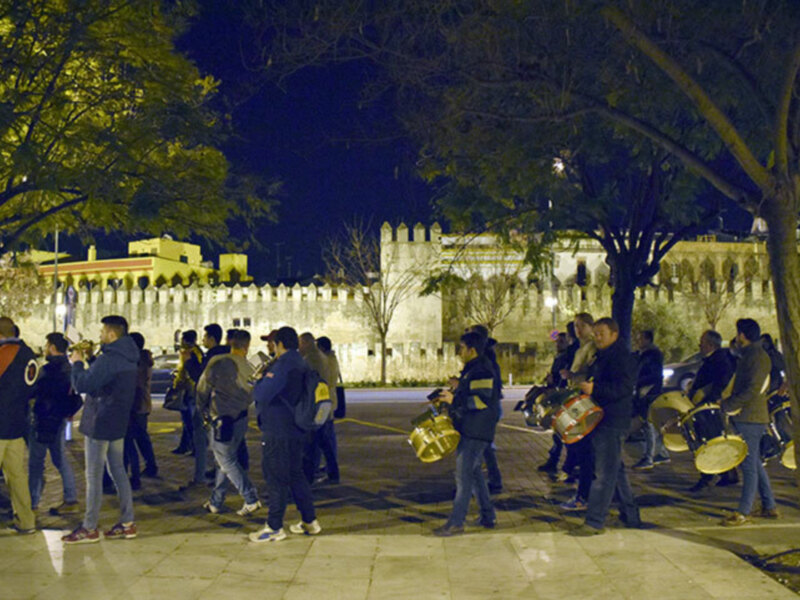Grupo de músicos tocando instrumentos en una plaza iluminada por la noche, con un castillo en el fondo.