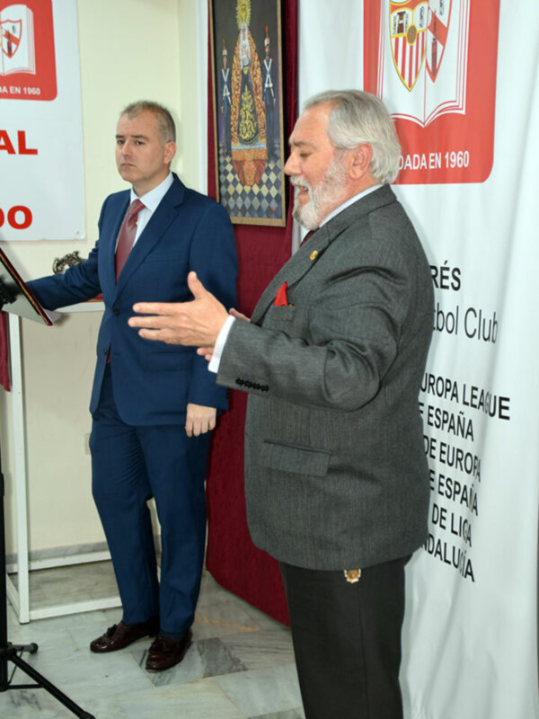 Dos hombres en trajes hablando en un evento formal, con banderas y carteles de fútbol en el fondo.