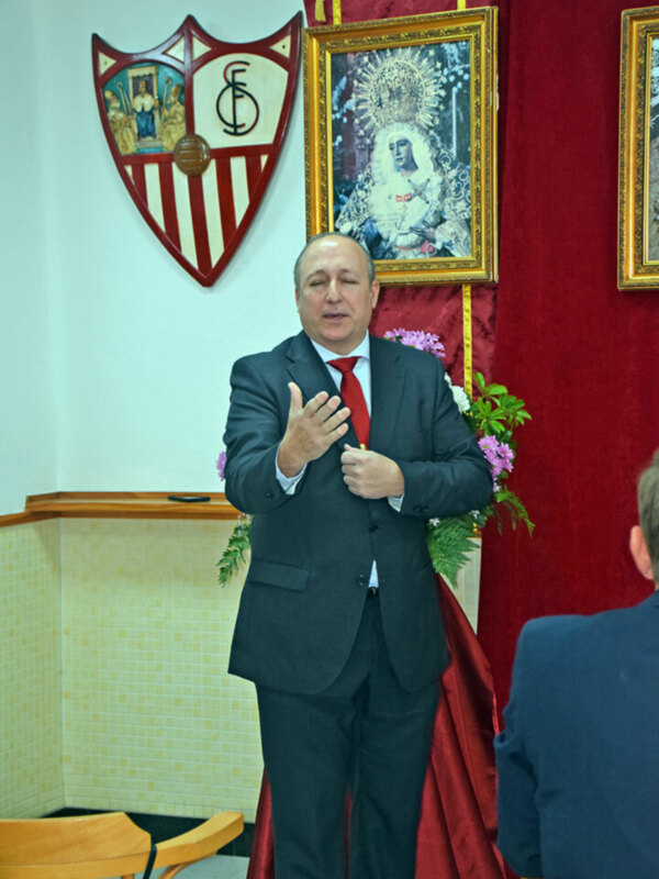 Un hombre en un traje negro hablando en una sala con un escudo y una imagen religiosa en el fondo.