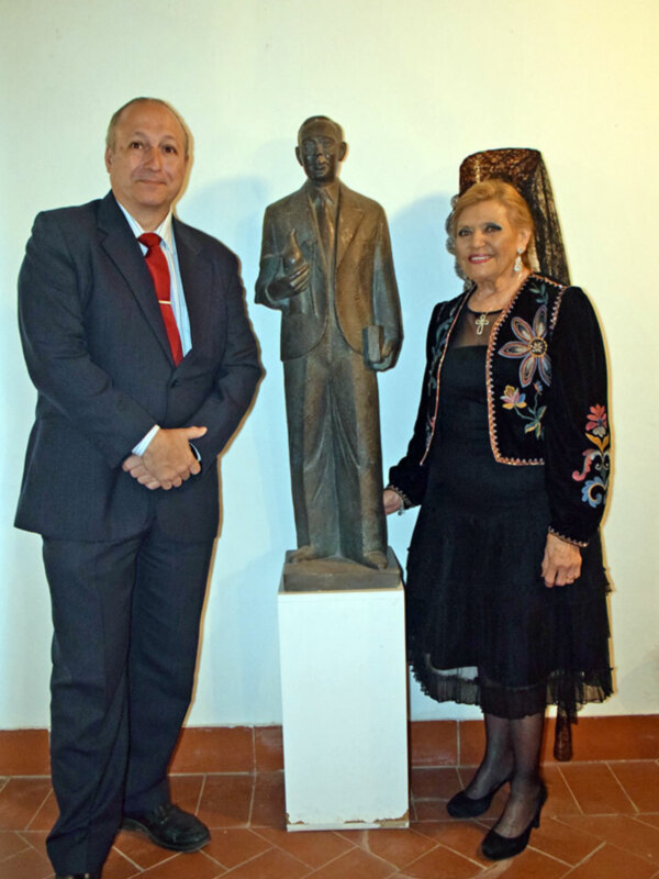 Dos personas posan junto a una estatua de bronce de un hombre en un espacio interior.