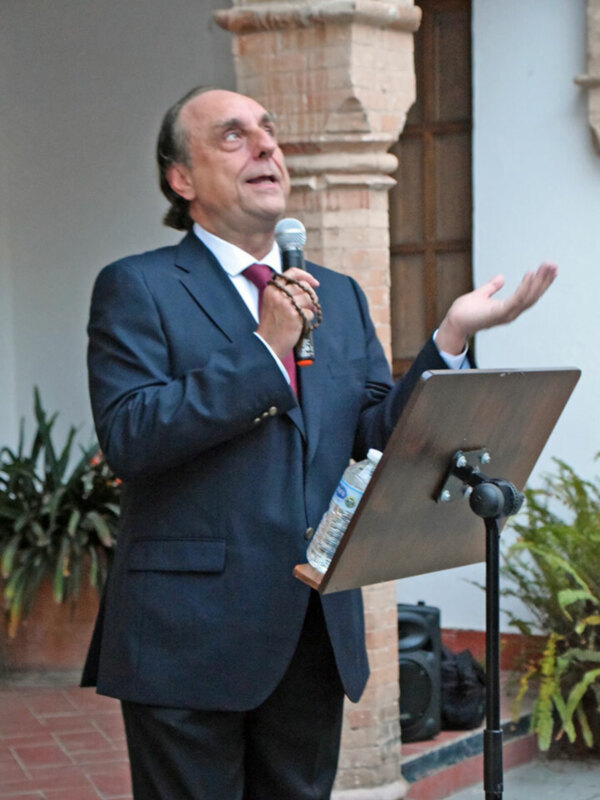 Hombre en traje hablando en un evento público con un micrófono y un soporte de música en el fondo.