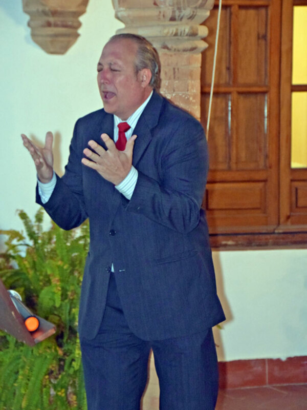 Hombre en traje hablando con expresión entusiasta en un entorno interior.