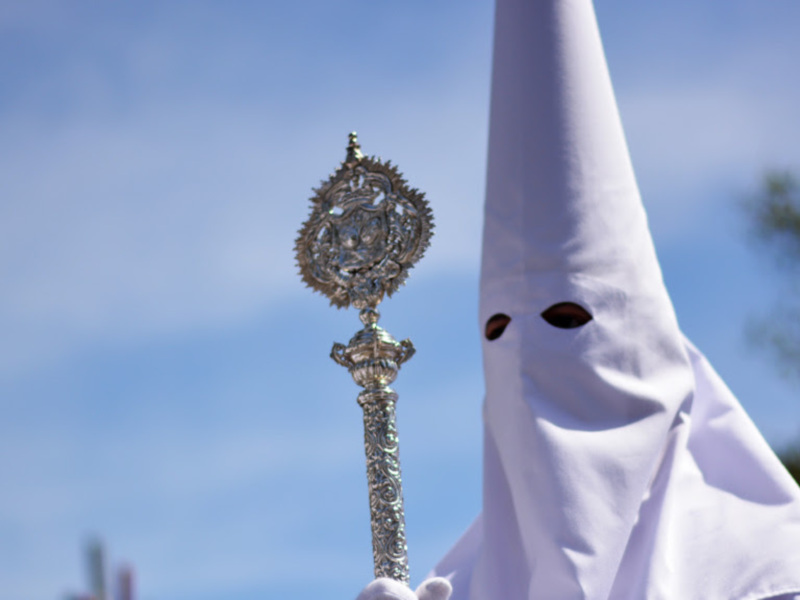 Procesión de Semana Santa con figura en capa blanca y manto ornado, bajo un cielo azul claro.