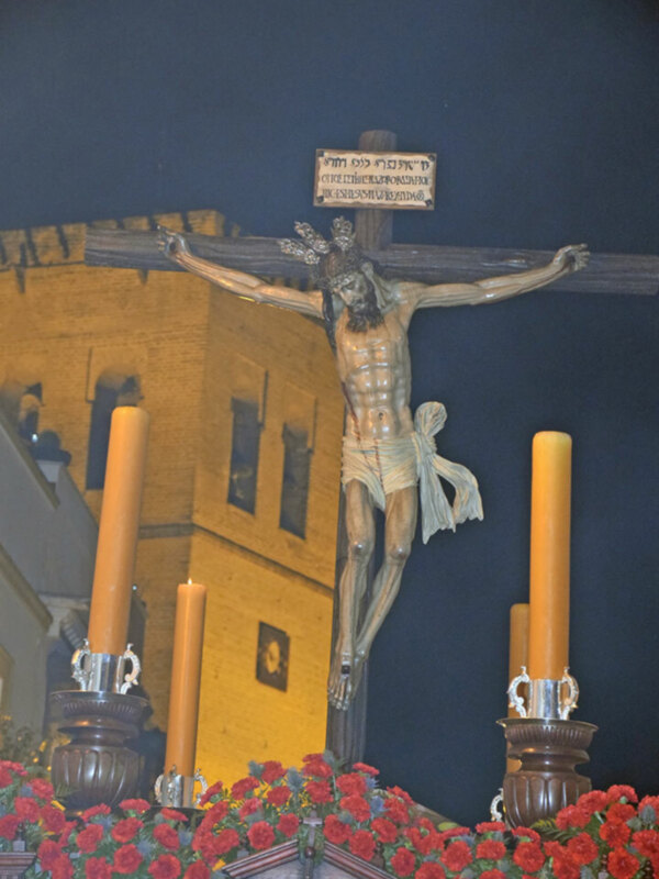 Crucificado en una iglesia con flores y velas, iluminado por la luz del exterior.