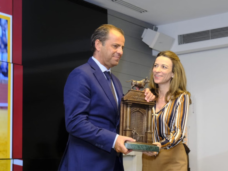 Entrega de un trofeo en un evento de Sevilla, con una pantalla en el fondo mostrando una imagen de una plaza de toros.