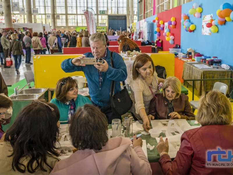 Una imagen de una feria o evento con personas tomando fotos y conversando en mesas con decoración colorida.