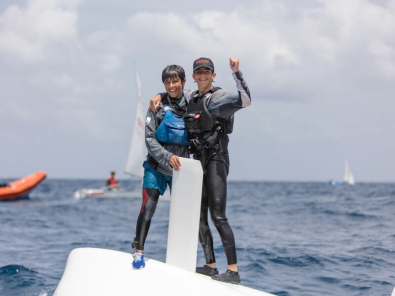Dos jóvenes celebran sobre un velero en el mar abierto, con equipos de seguridad y fondo de otros barcos navegando.
