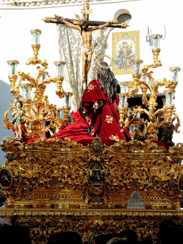 Procesión religiosa con estandarte y figura de Jesús en la cruz, adornada con detalles dorados y ornamentación.