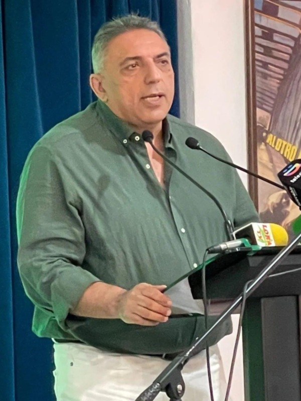 Hombre hablando en un evento público con micros de televisión en el escenario.