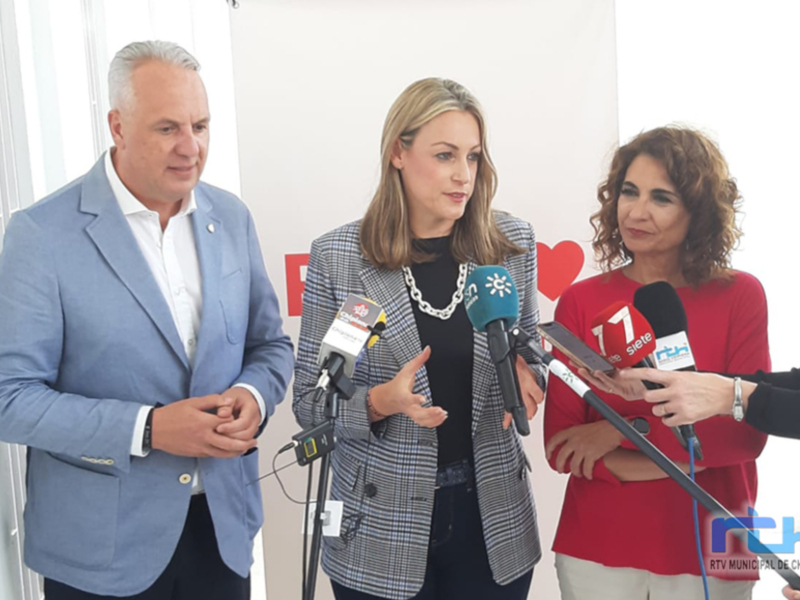 Una mujer habla en un micrófono mientras dos personas la escuchan, posiblemente en una conferencia de prensa.