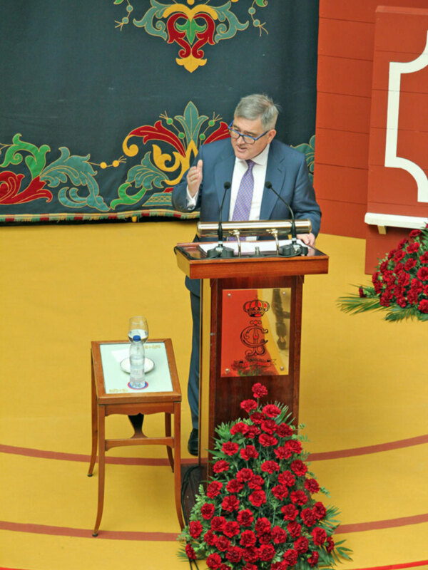 Un hombre habla desde un podio en un evento formal, con flores y decoración festiva al fondo.
