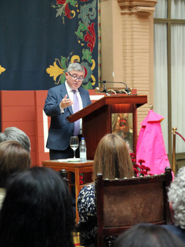 Speaker addressing an audience in a formal setting with a decorative backdrop and floral decorations on the podium.