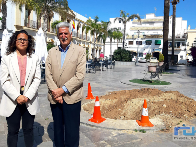 Dos personas posan frente a un área de obras en una plaza con palmeras y edificios blancos en Chipiona, España.