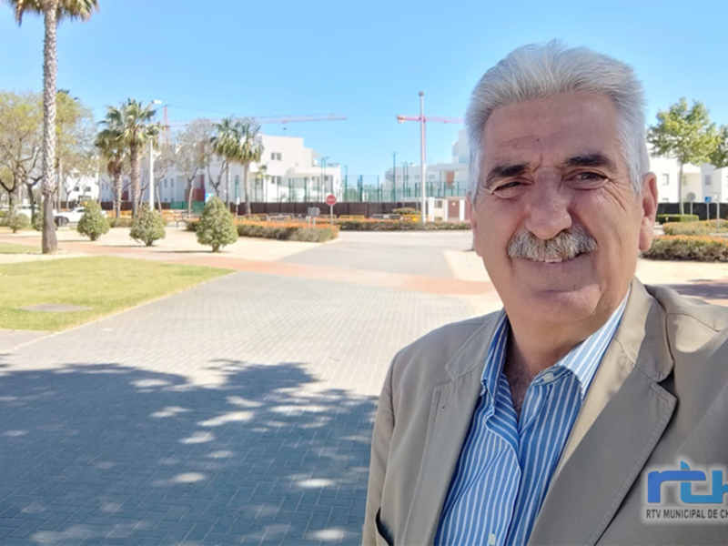 Retrato de un hombre en un parque con edificios y palmeras al fondo, bajo un cielo azul claro.