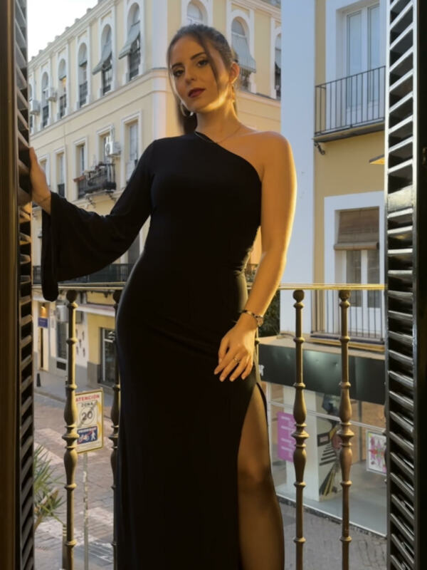 Elegante mujer en vestido negro con abertura lateral, posando en un balcón con edificios históricos de fondo.