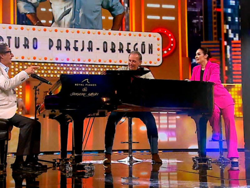 Un piano en el escenario de un programa de televisión, con dos personas sentadas en él. El piano es de la marca Royal Pianos, y el fondo tiene un diseño de luces y letras en el escenario.