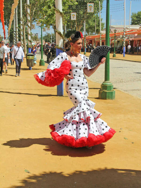 Una mujer vestida con un traje de flamenca baila en una calle con banderolas y palmeras en el fondo.