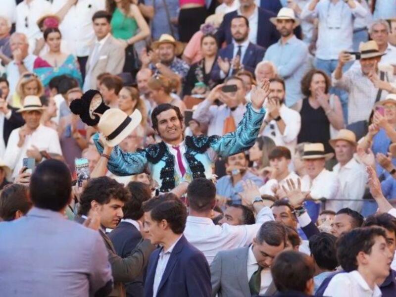 Una celebración en un evento público, con una figura en traje y sombrero de copa, rodeada de personas en trajes formales y sombreros de copa, capturando la atención de la multitud con su gesto de saludo.