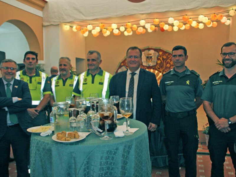 Grupo de personas en uniforme junto a un hombre en traje, posando para una foto en una reunión formal. La mesa está adornada con copas de vino y una toalla verde. La iluminación es cálida con luces colgantes en el fondo.