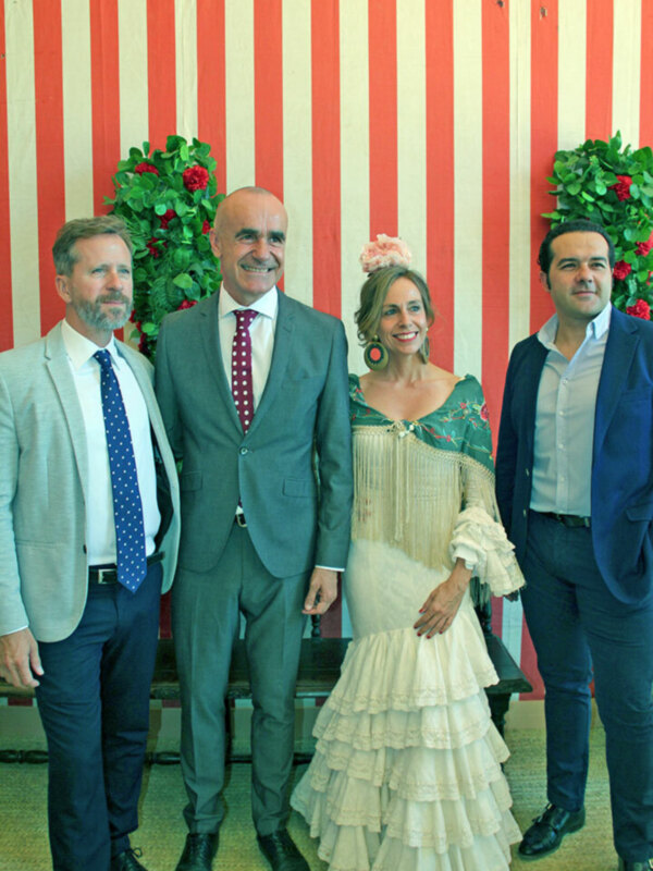Tres personas posando frente a un fondo con rayas rojas y blancas, uno de ellos con un vestido blanco con flecos y detalles florales, y otro con una camisa blanca y chaqueta azul.
