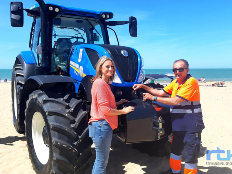 Dos personas posan junto a un tractor azul en una playa con el mar al fondo.