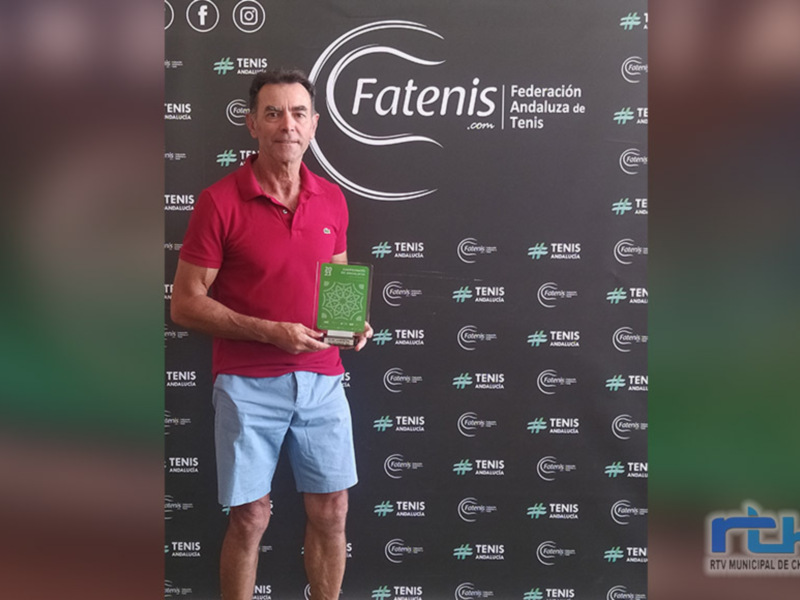 Persona con camiseta roja y shorts azules posando con un trofeo en frente de un fondo con logos de la Federación Andaluza de Tenis.