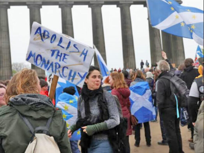 Manifestación con pancarta 'Abolish the Monarchy' y banderas de Escocia y la UE en un monumento con columnas.