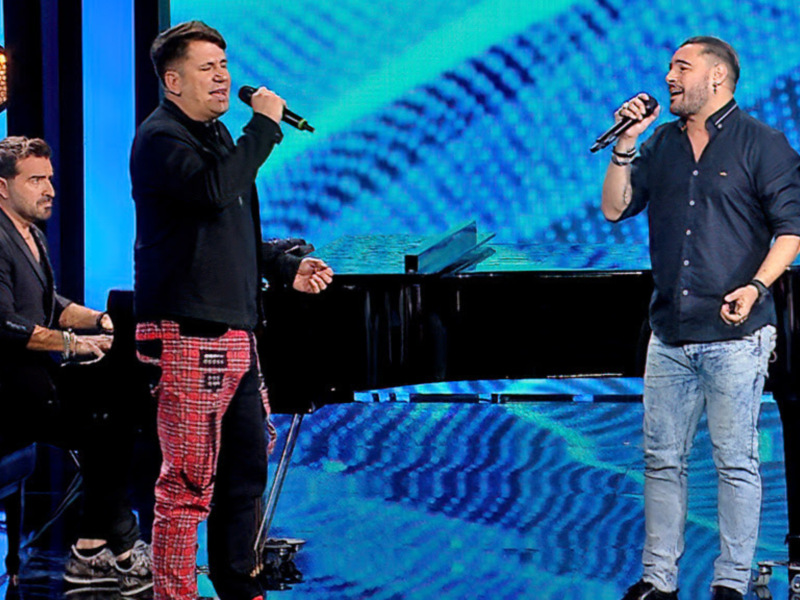 Tres artistas en escenario con piano, uno tocando y otros cantando, bajo luces y fondo azul.