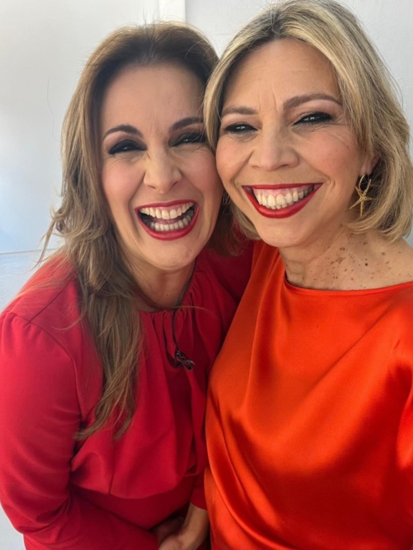 Dos personas sonrientes, posando juntas en una foto selfie, ambos con vestidos rojos y maquillaje brillante, posando frente a una pared blanca.