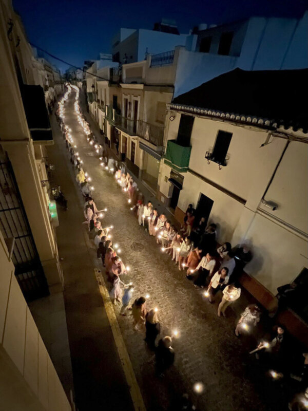 Procesión nocturna con velas en una calle adoquinada, iluminada por la luz de las velas y las fachadas de edificios blancos.