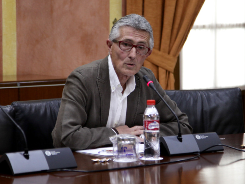 Un hombre con gafas y una chaqueta marrón se sienta en una sala de conferencias, frente a una mesa con un microfono y una botella de agua. La pared detrás de él tiene paneles de colores y cortinas.