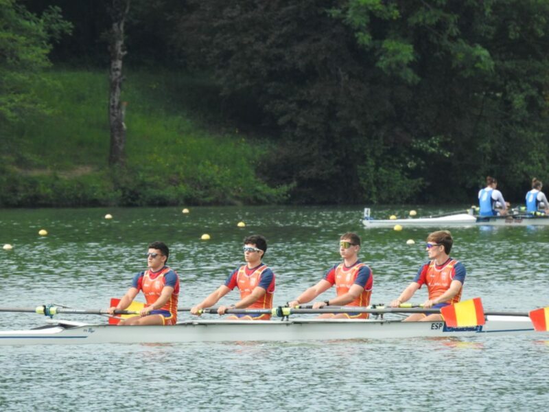 Equipo de remo en competición, luciendo uniformes naranjas y azules, remando en un lago con árboles al fondo.