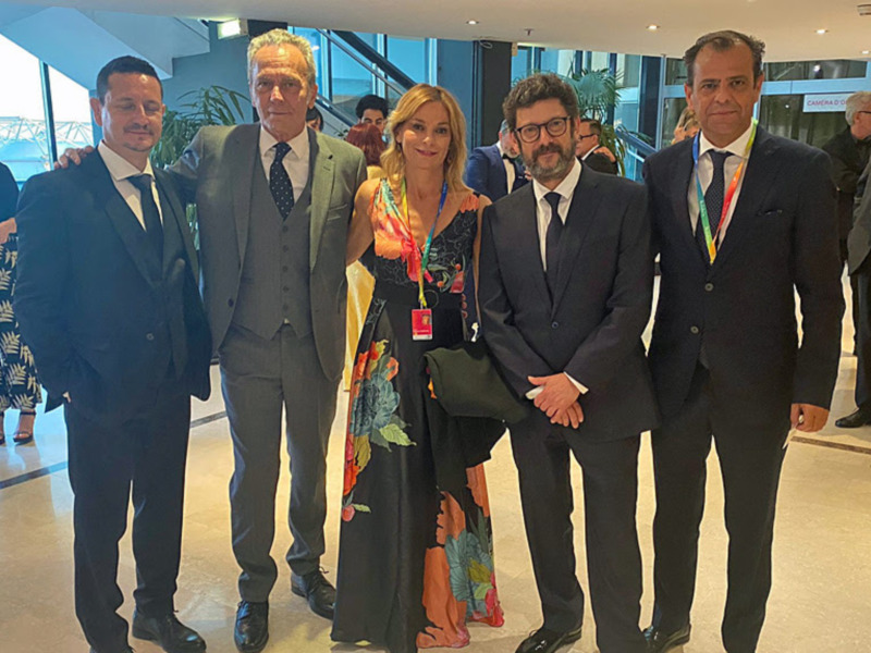 Grupo de personas en un evento formal, posando para una foto en un entorno moderno con ventanas y plantas.