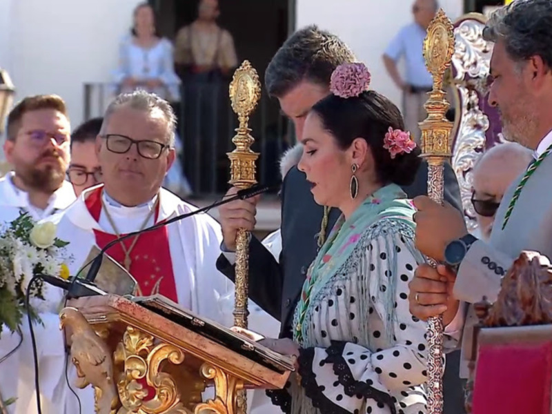 Celebración religiosa con un sacerdote y una mujer vestida con traje típico andaluz en un entorno festivo.