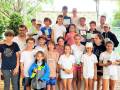Un grupo de niños y jóvenes posan orgullosos con sus trofeos en un torneo de tenis. La imagen muestra a los participantes sosteniendo sus premios, con un fondo verde y una bandera en el cielo.